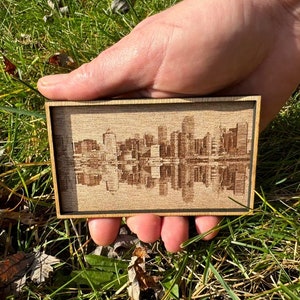 May include: A small wooden plaque with a laser-engraved cityscape design. The plaque is brown with a light wood grain and has a dark brown border.