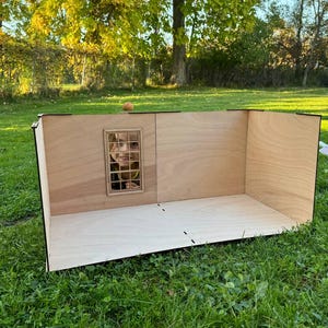 May include: A light-colored wooden dollhouse room structure with a flat base and three walls. One wall features a rectangular window frame with a grid pattern. The structure is constructed from thin plywood panels with visible dark edges.
