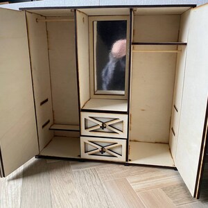 May include: A light-colored wooden dollhouse wardrobe with two open doors, a central mirror, and two small drawers. The interior features a hanging rod on the left and right sides, with a shelf above the drawers. The wood edges are dark brown.