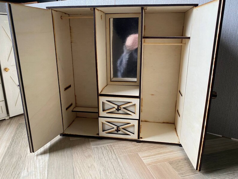 May include: A light-colored wooden dollhouse wardrobe with two open doors, a central mirror, and two small drawers. The interior features a hanging rod on the left and right sides, with a shelf above the drawers. The wood edges are dark brown.