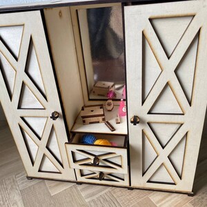 May include: A light-colored wooden doll wardrobe featuring two hinged doors with X-shaped cutouts, a central mirror, and two stacked drawers. The interior contains small wooden accessories, including a box and miniature items, with dark edges.