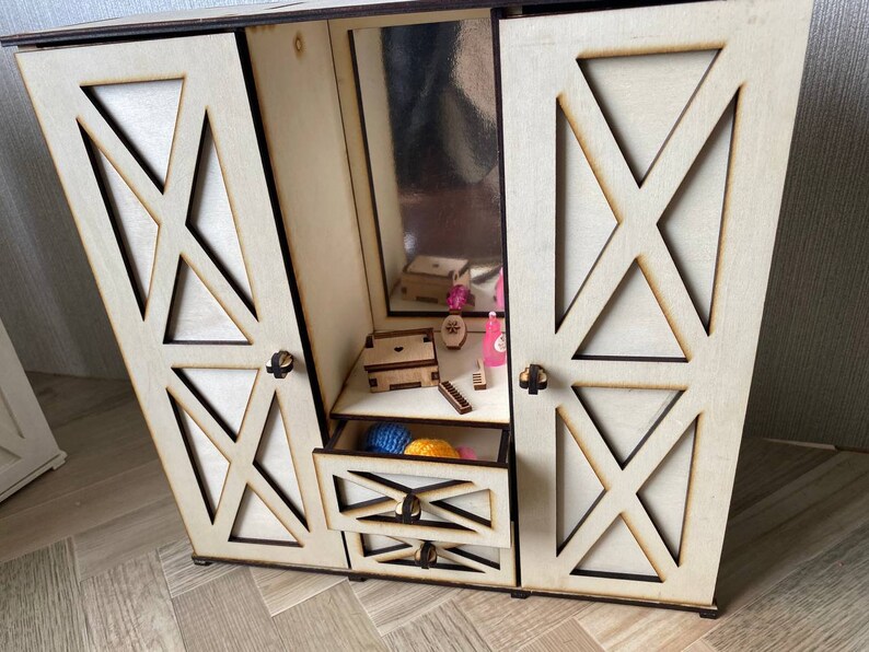 May include: A light-colored wooden doll wardrobe featuring two hinged doors with X-shaped cutouts, a central mirror, and two stacked drawers. The interior contains small wooden accessories, including a box and miniature items, with dark edges.