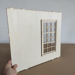 May include: A flat, rectangular light-colored wooden panel featuring a cutout window frame with a grid pattern. The panel has small tabs along the top and bottom edges, indicating it is a component designed for assembly into a larger structure.
