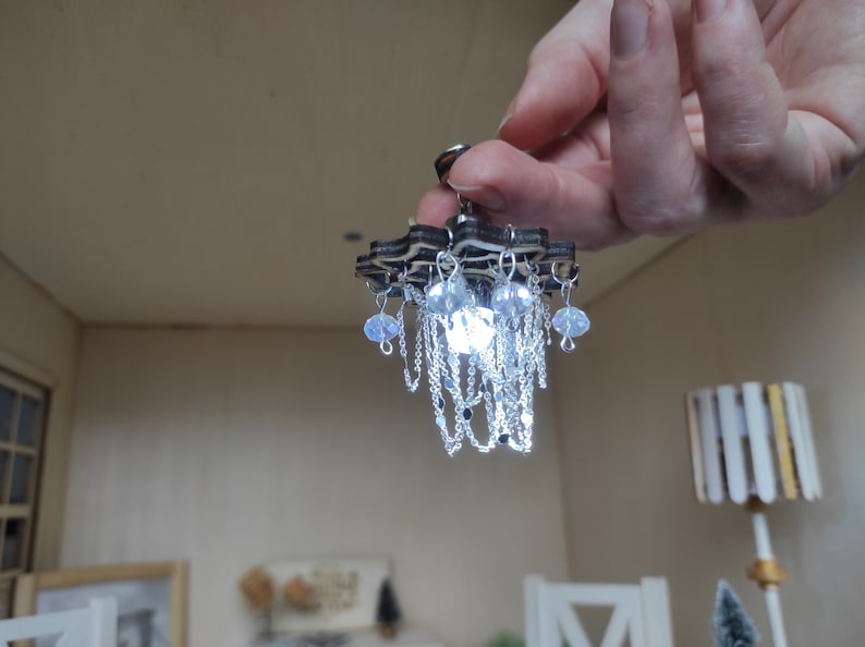 May include: A miniature wooden chandelier with a star-shaped base and crystal-like drops hanging from it. The chandelier is lit up with a white light.