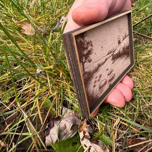 May include: A small wooden frame with a laser-engraved image of a river scene. The frame is made of dark wood and has a light wood interior. The image shows a river flowing through a field with trees on either side. There are rocks in the river.