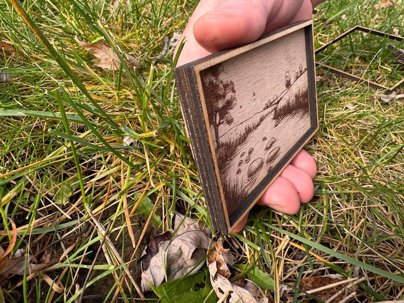 May include: A small wooden frame with a laser-engraved image of a river scene. The frame is made of dark wood and has a light wood interior. The image shows a river flowing through a field with trees on either side. There are rocks in the river.