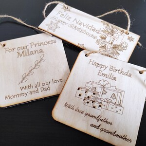 May include: Three wooden ornaments with twine hangers. The first ornament says "Feliz Navidad Merry Christmas" with a pine bough and candles. The second ornament says "For our Princess Milana" with a small branch. The third ornament says "Happy Birthday Emilia" with a stack of wrapped presents.