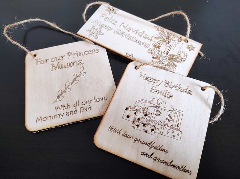 May include: Three wooden ornaments with twine hangers. The first ornament says "Feliz Navidad Merry Christmas" with a pine bough and candles. The second ornament says "For our Princess Milana" with a small branch. The third ornament says "Happy Birthday Emilia" with a stack of wrapped presents.