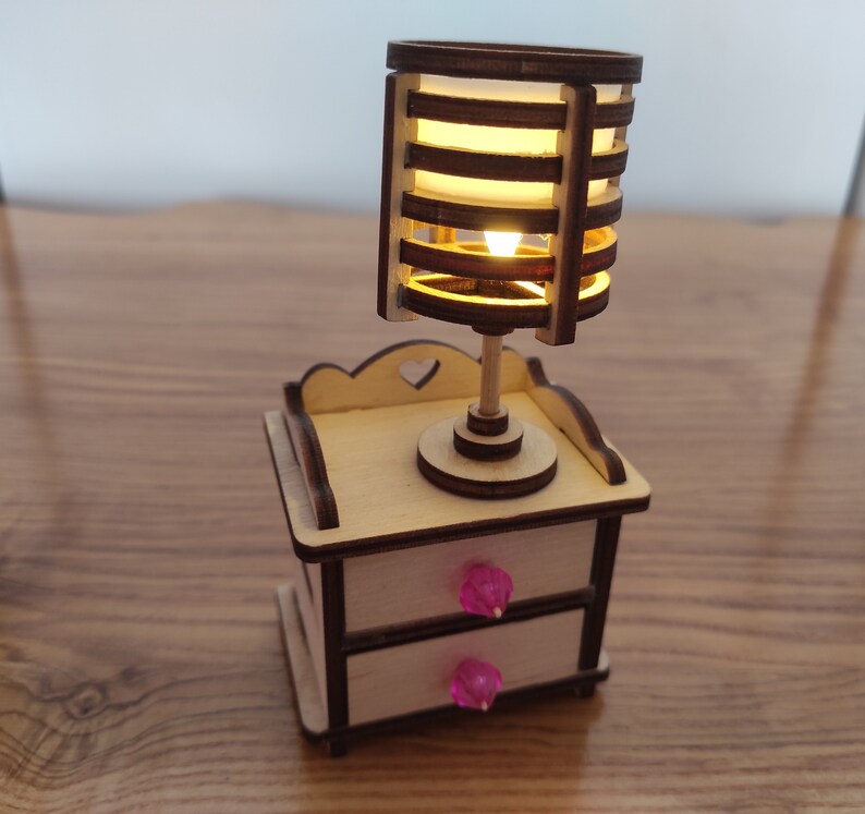 May include: A wooden nightstand with two drawers and a lamp with a slatted shade. The drawers have pink knobs and the lamp is lit.