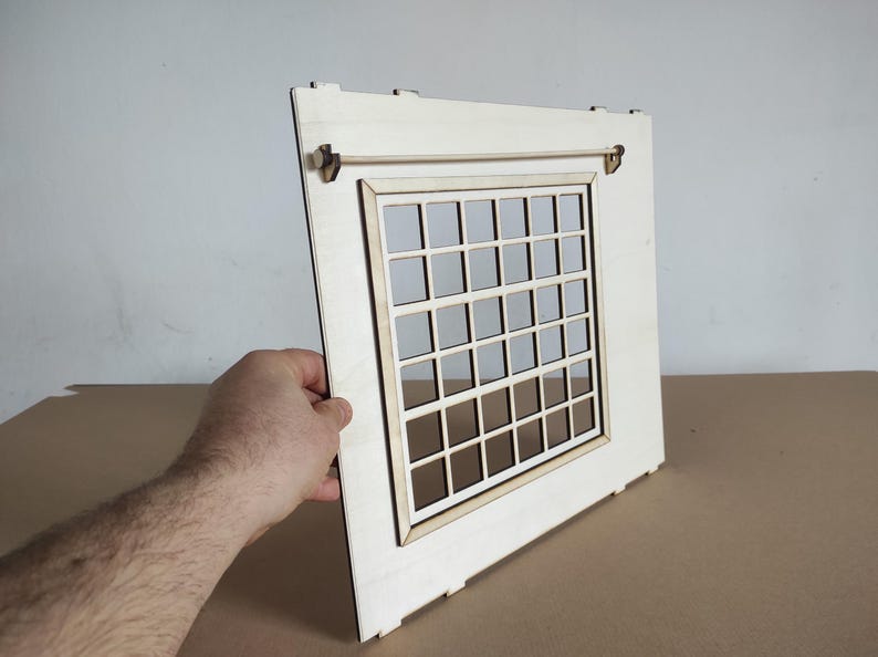May include: A light-colored wooden panel featuring a rectangular window cutout with a grid pattern. The panel includes a horizontal rod mounted near the top edge with small brackets, designed for assembly as part of a larger structure or model.
