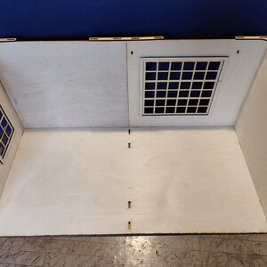 May include: A white wooden dollhouse room with two windows, one on each side. The windows have black frames and are made of a grid pattern. The room is empty and has a white wooden floor.