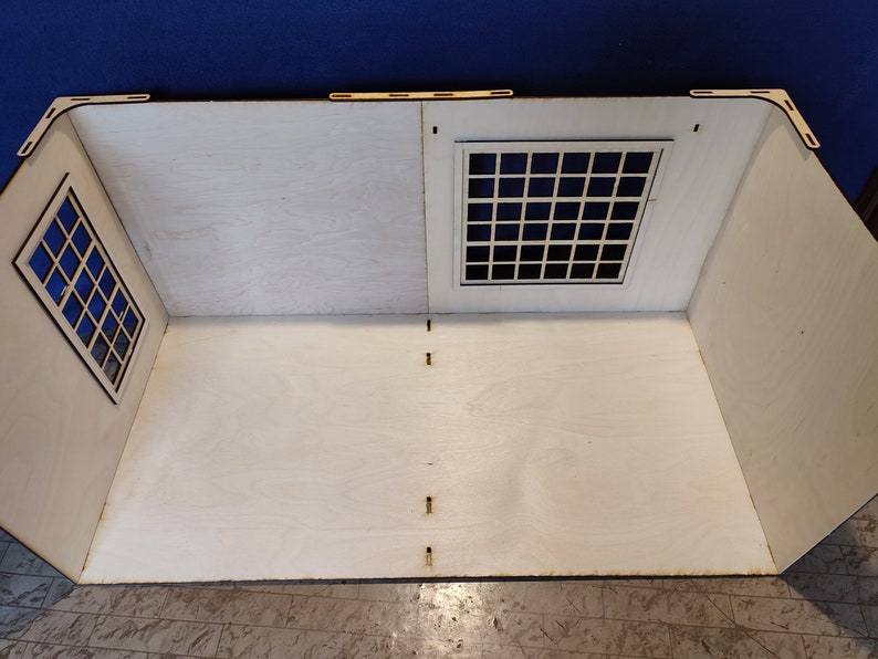 May include: A white wooden dollhouse room with two windows, one on each side. The windows have black frames and are made of a grid pattern. The room is empty and has a white wooden floor.