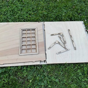 May include: A wooden model kit with a window and several pieces of wood for assembling the structure. The pieces are laid out on a light brown surface.