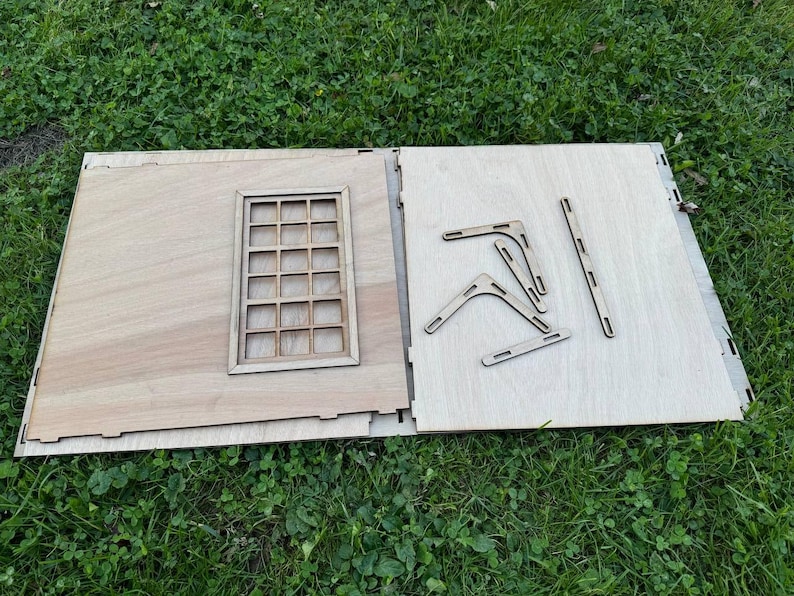 May include: A wooden model kit with a window and several pieces of wood for assembling the structure. The pieces are laid out on a light brown surface.