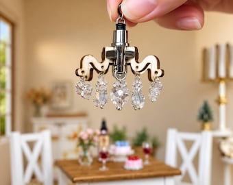 May include: A miniature chandelier ornament featuring a central silver-toned metal post, light-colored wooden arms with a wavy design, and multiple clear, faceted crystal-like beads hanging from the structure, suspended by a small metal jump ring.