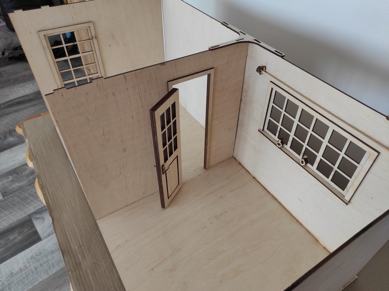 May include: A wooden dollhouse room with two windows and a door. The walls are made of light wood and the floor is made of a darker wood.