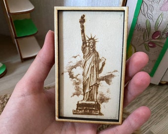 May include: A wooden frame with a laser-engraved image of the Statue of Liberty. The image is in shades of brown and white, and the frame is a light brown color.