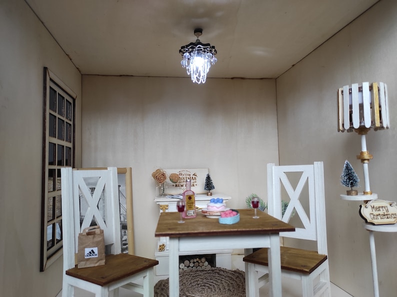 May include: A miniature dollhouse dining room with a white table and chairs, a small chandelier, a bottle of wine, a cake, and a Christmas tree. The room is decorated with a sign that says "Wishing you a Merry Christmas and a Happy New Year".
