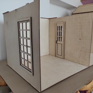 May include: A wooden dollhouse room with a window and a door. The walls are made of plywood and the floor is unfinished.