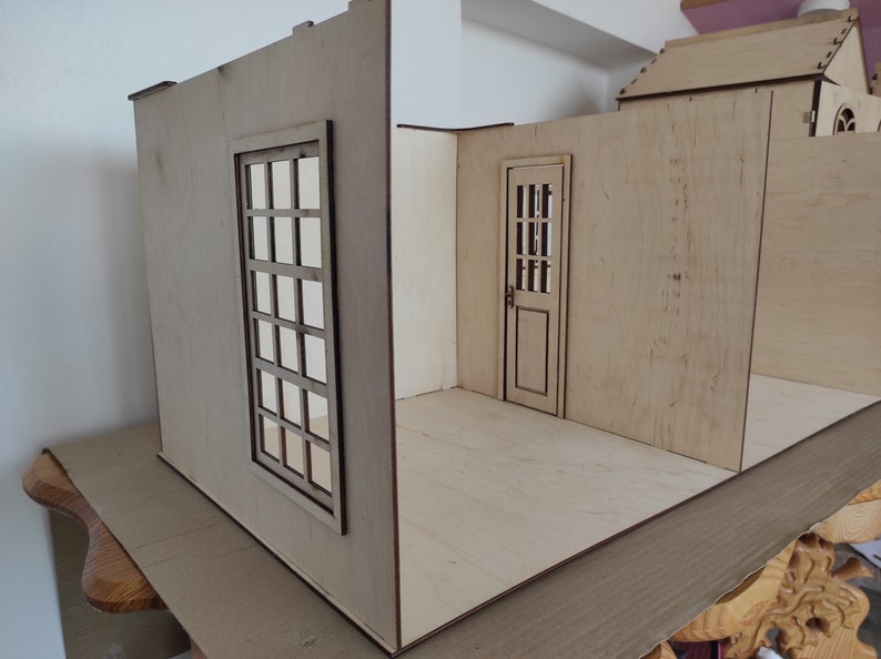 May include: A wooden dollhouse room with a window and a door. The walls are made of plywood and the floor is unfinished.