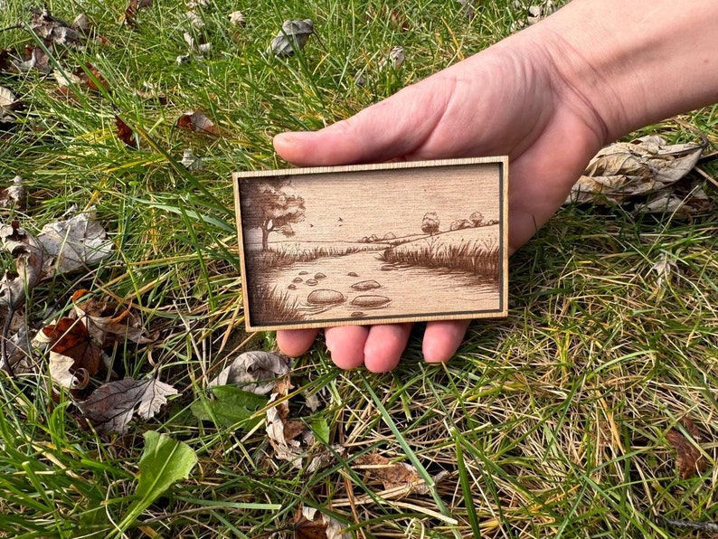 May include: A small wooden plaque with a laser-engraved image of a river scene. The scene features a winding river with trees and rocks along the banks. The plaque is held in a hand.