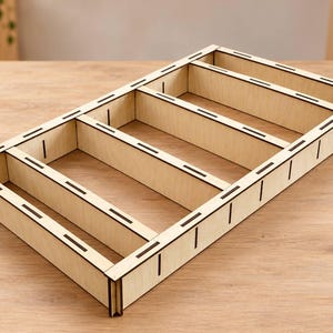 May include: A rectangular light-colored wooden organizer tray featuring four open compartments. The structure is constructed from thin, flat panels with visible laser-cut slots and joints along the edges, creating a grid-like storage frame.