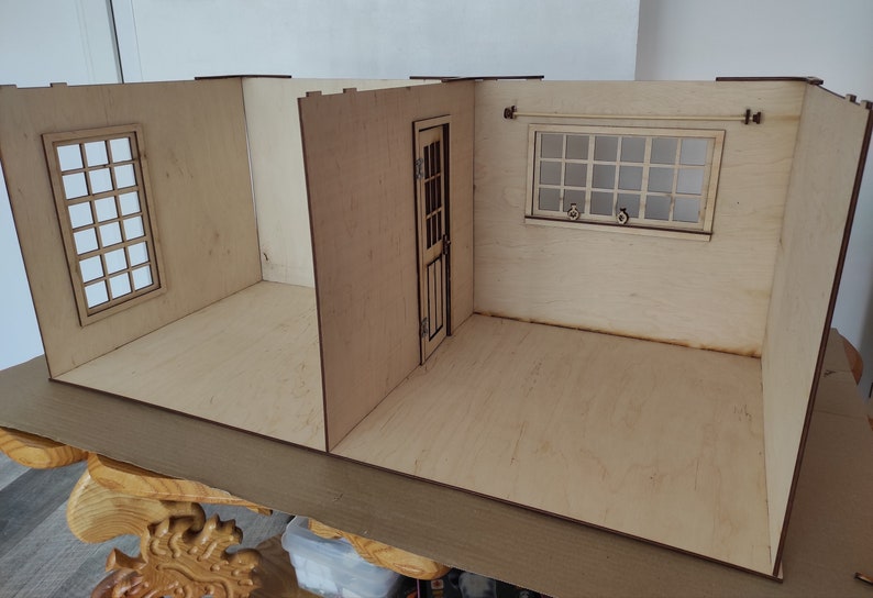 May include: A wooden dollhouse with three rooms, one with a window, one with a door, and one with a window and a curtain rail. The dollhouse is made of light wood and has a natural finish.