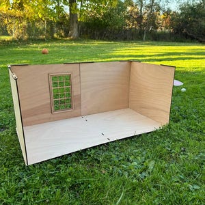 May include: A light-colored wooden dollhouse room structure featuring a flat floor and two connected walls. One wall includes a rectangular window frame with a grid pattern. The structure is constructed from thin plywood panels with visible joints.