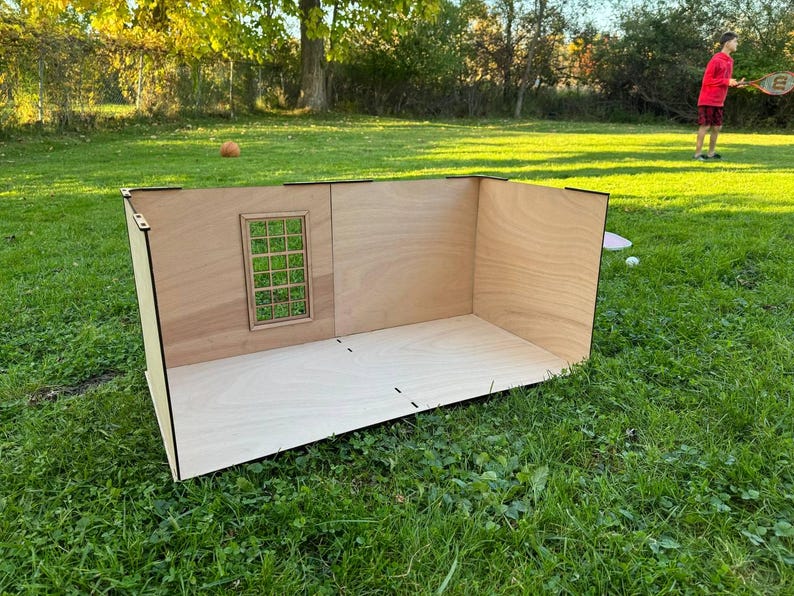 May include: A light-colored wooden dollhouse room structure featuring a flat floor and two connected walls. One wall includes a rectangular window frame with a grid pattern. The structure is constructed from thin plywood panels with visible joints.