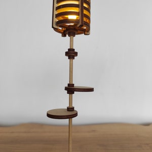 May include: A wooden lamp with a light brown finish and a geometric design. The lamp has a cylindrical shade with a light bulb inside. The lamp has two shelves, one above the other, and a wooden pole that supports the shade and shelves.