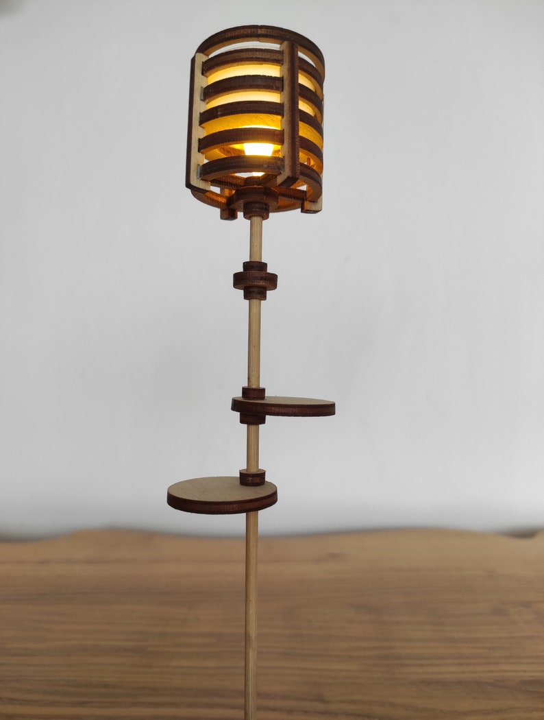 May include: A wooden lamp with a light brown finish and a geometric design. The lamp has a cylindrical shade with a light bulb inside. The lamp has two shelves, one above the other, and a wooden pole that supports the shade and shelves.