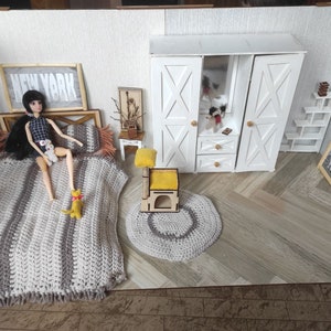 May include: A dollhouse bedroom with a doll lying on a bed with a crocheted blanket. The room has a white wardrobe with a mirror, a small side table, a cat tree, and a rug. The walls are white with a framed print that says "NEW YORK".