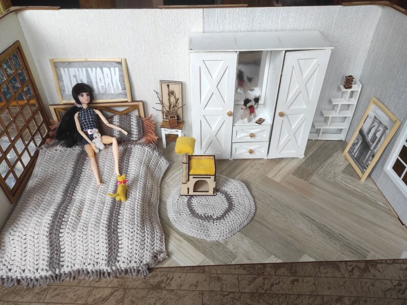 May include: A dollhouse bedroom with a doll lying on a bed with a crocheted blanket. The room has a white wardrobe with a mirror, a small side table, a cat tree, and a rug. The walls are white with a framed print that says "NEW YORK".