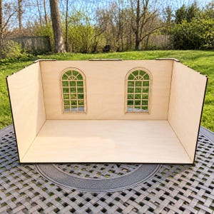 May include: A light-colored wooden dollhouse room structure featuring a flat floor, two side walls, and a back wall with two arched windows. The windows have a grid pattern and a semi-circular top section. The structure is made of thin plywood.