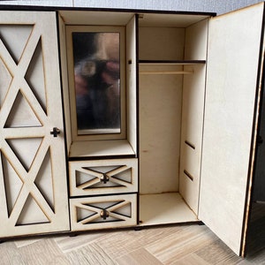 May include: A light-colored wooden doll wardrobe featuring a left door with an X-shaped cutout design, a central vertical mirror above two small drawers with matching X-shaped accents, and an open right section with a horizontal hanging rod.