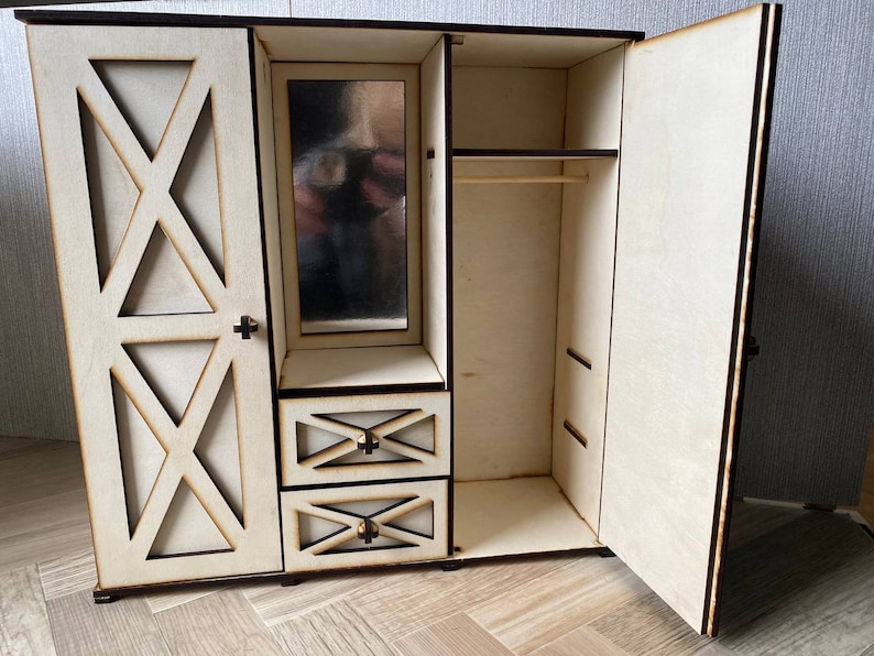 May include: A light-colored wooden doll wardrobe featuring a left door with an X-shaped cutout design, a central vertical mirror above two small drawers with matching X-shaped accents, and an open right section with a horizontal hanging rod.