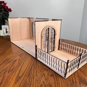 May include: A miniature wooden balcony with a detailed balustrade and arched doorway. The structure is made of light-colored wood with black accents, featuring a small window and double doors. The balcony is designed for a dollhouse or similar craft project.