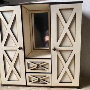 May include: A light-colored wooden dollhouse wardrobe featuring two side doors with X-shaped cutouts, a central vertical mirror, and two stacked drawers below the mirror. Each door and drawer is equipped with a small, dark-colored knob handle.