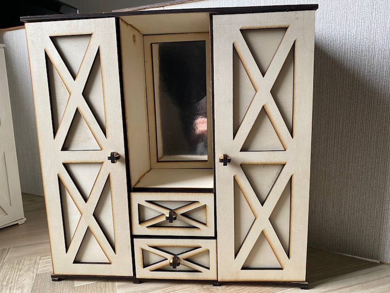 May include: A light-colored wooden dollhouse wardrobe featuring two side doors with X-shaped cutouts, a central vertical mirror, and two stacked drawers below the mirror. Each door and drawer is equipped with a small, dark-colored knob handle.