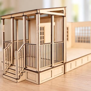 Könnte beinhalten: Ein helles Holzmodellhaus-Set mit offenem Design, einer Veranda, vertikalen Geländern und einer Treppe. Die Struktur umfasst ein Dach, Stützpfeiler und einen Fensterrahmen mit Gittermuster an der seitlichen Wandsektion.