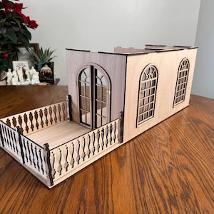 May include: A miniature wooden building with a balcony and arched windows. The building is made of light-colored wood with dark brown accents. The balcony has a decorative railing. The windows have a detailed grid pattern.