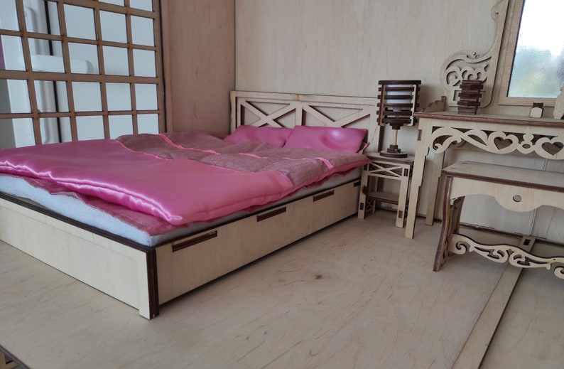 May include: A wooden dollhouse bedroom set with a pink bed, a wooden nightstand, and a vanity with a mirror. The bed has a pink satin comforter and a wooden headboard.