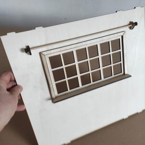 May include: A light-colored wooden wall panel featuring a rectangular window with a grid of sixteen panes. A thin horizontal rod is mounted above the window frame with small dark brackets on each side. The panel has notched edges for assembly.
