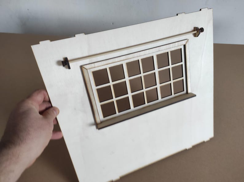 May include: A light-colored wooden wall panel featuring a rectangular window with a grid of sixteen panes. A thin horizontal rod is mounted above the window frame with small dark brackets on each side. The panel has notched edges for assembly.