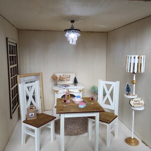May include: A miniature dollhouse dining room with a white table and four chairs, a small chandelier, a bottle of wine, a cake, and a floor lamp with a wooden shade. The walls are beige and the floor is white. The lamp has a sign that says "Merry Christmas".