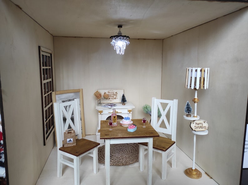 May include: A miniature dollhouse dining room with a white table and four chairs, a small chandelier, a bottle of wine, a cake, and a floor lamp with a wooden shade. The walls are beige and the floor is white. The lamp has a sign that says "Merry Christmas".