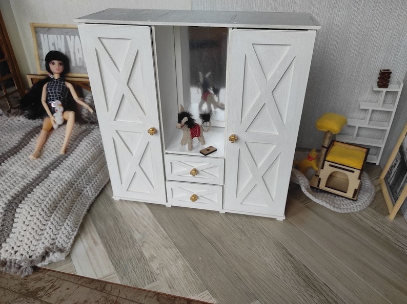 May include: A white wooden dollhouse wardrobe featuring two side doors with X-shaped paneling, a central open section with a mirror, and two small drawers below. The unit is accented with small gold-toned round knobs on the doors and drawers.
