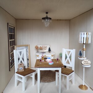 May include: A miniature dollhouse dining room with a white table and four chairs, a small chandelier, a framed picture, a small Christmas tree, and a floor lamp with a wooden shade and a sign that says "Merry Christmas".
