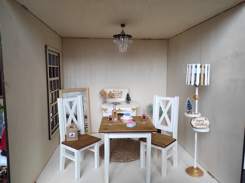 May include: A miniature dollhouse dining room with a white table and four chairs, a small chandelier, a framed picture, a small Christmas tree, and a floor lamp with a wooden shade and a sign that says "Merry Christmas".
