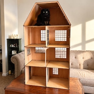 May include: A tall, multi-story wooden dollhouse with a peaked roof and open front design. The structure features multiple rooms with grid-patterned windows on the walls. A black cat is sitting inside the top attic level of the wooden dollhouse.